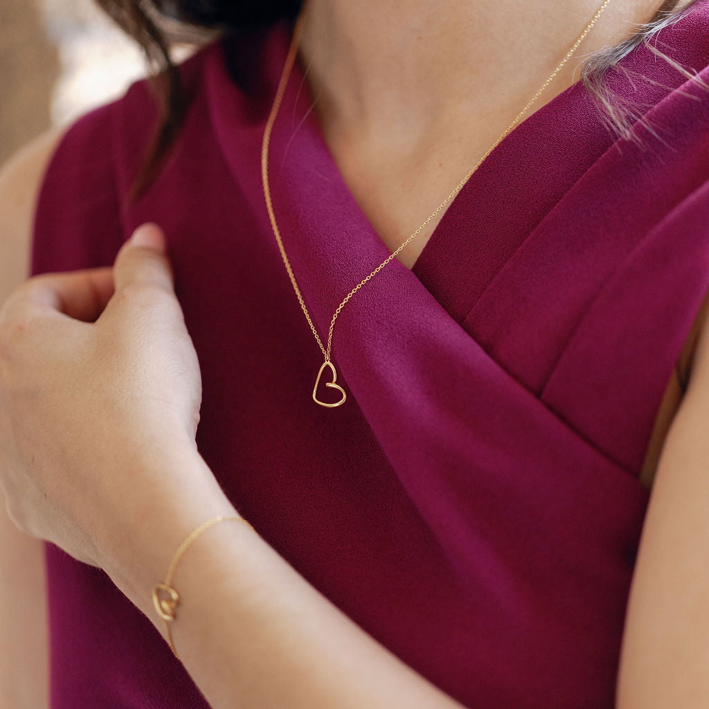 Person wearing a gold heart-shaped necklace and bracelet against a blurred background