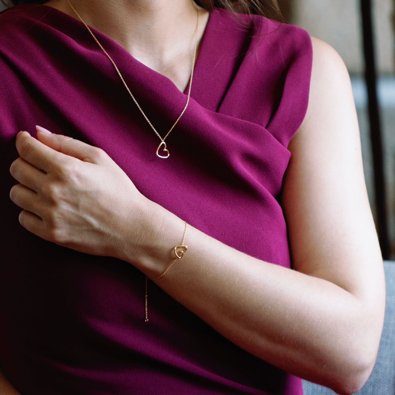 Person wearing a gold heart-shaped necklace and bracelet against a blurred background