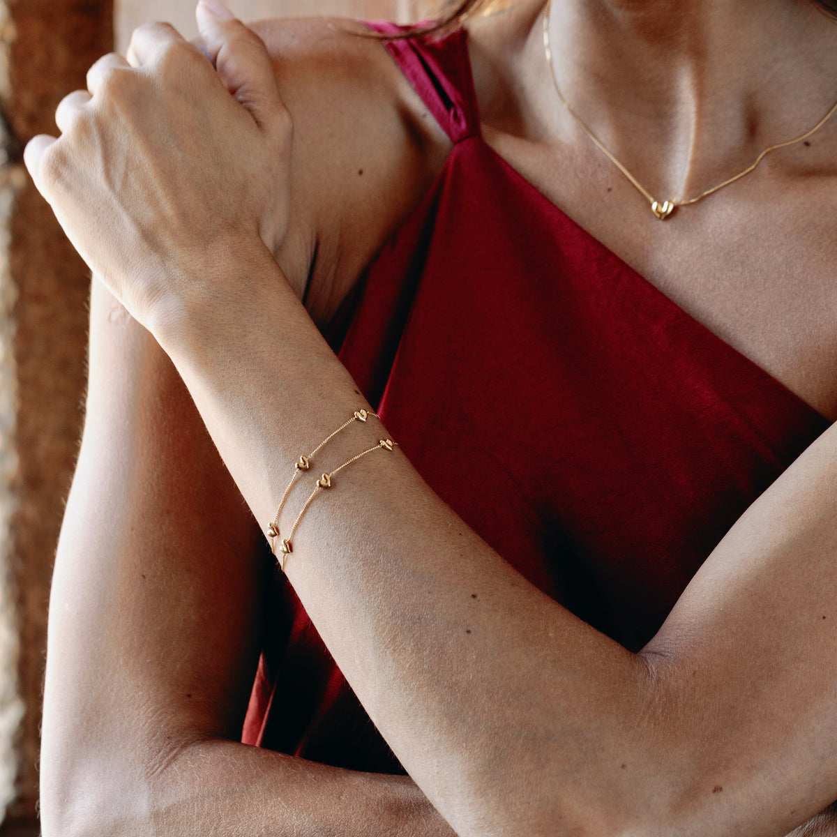 Close-up of a person wearing a gold vermeil bracelet and necklace with heart shaped pendants, with a blurred background.